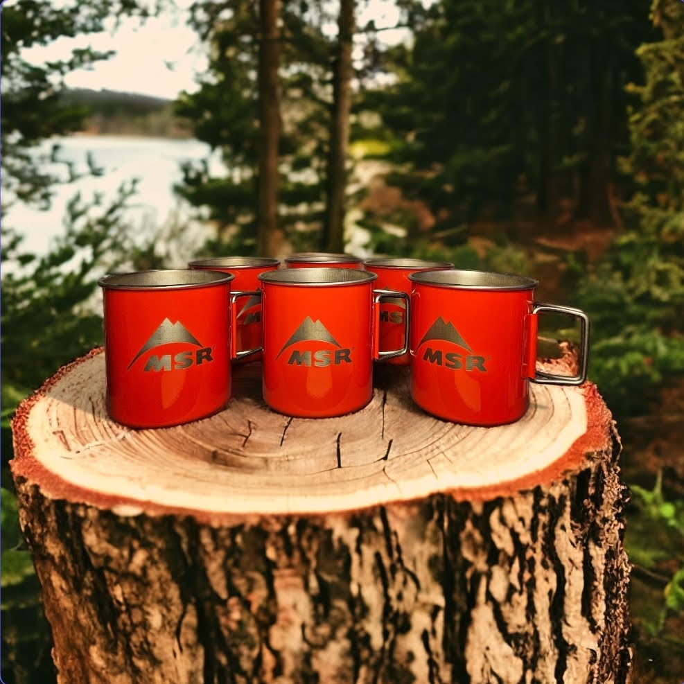 Laser-engraved red camping mugs branded with the MSR logo displayed on a wood stump in a forest setting.  Made by Green Fern Studio, based in Seattle.