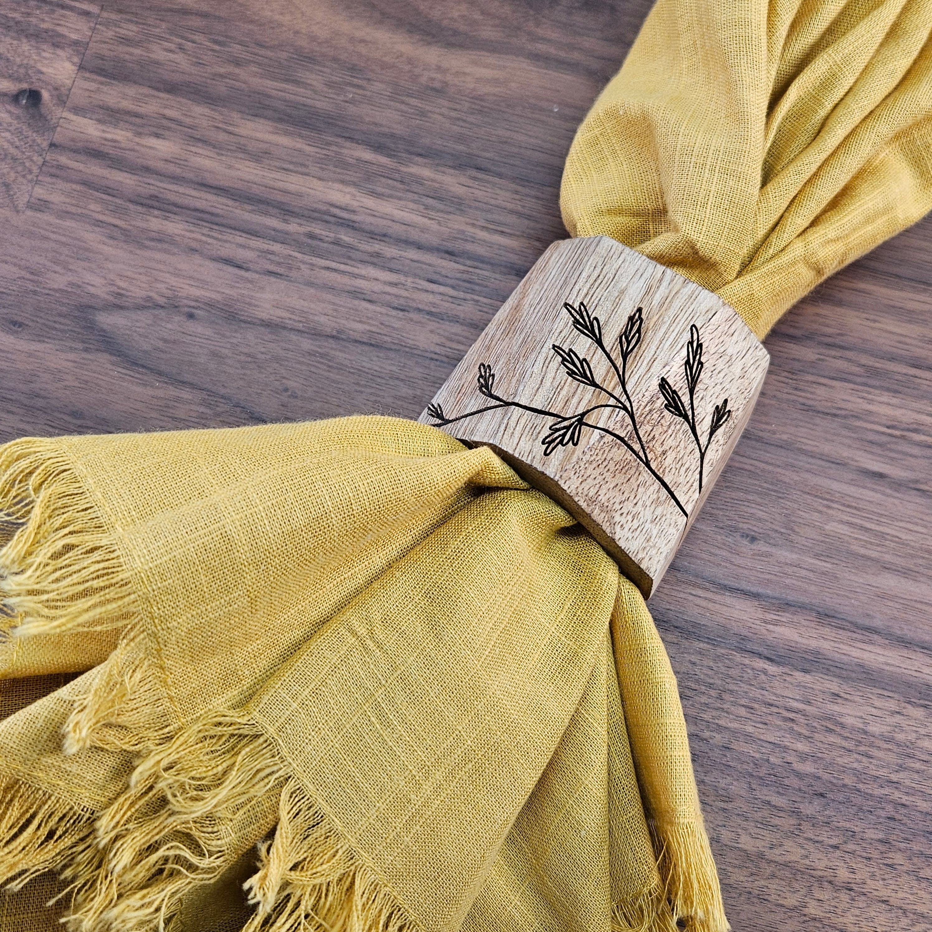 Laser engraved light wood napkin ring with delicate floral design holding a yellow napkin on a wood table surface. Made by Green Fern Studio, based in Seattle.