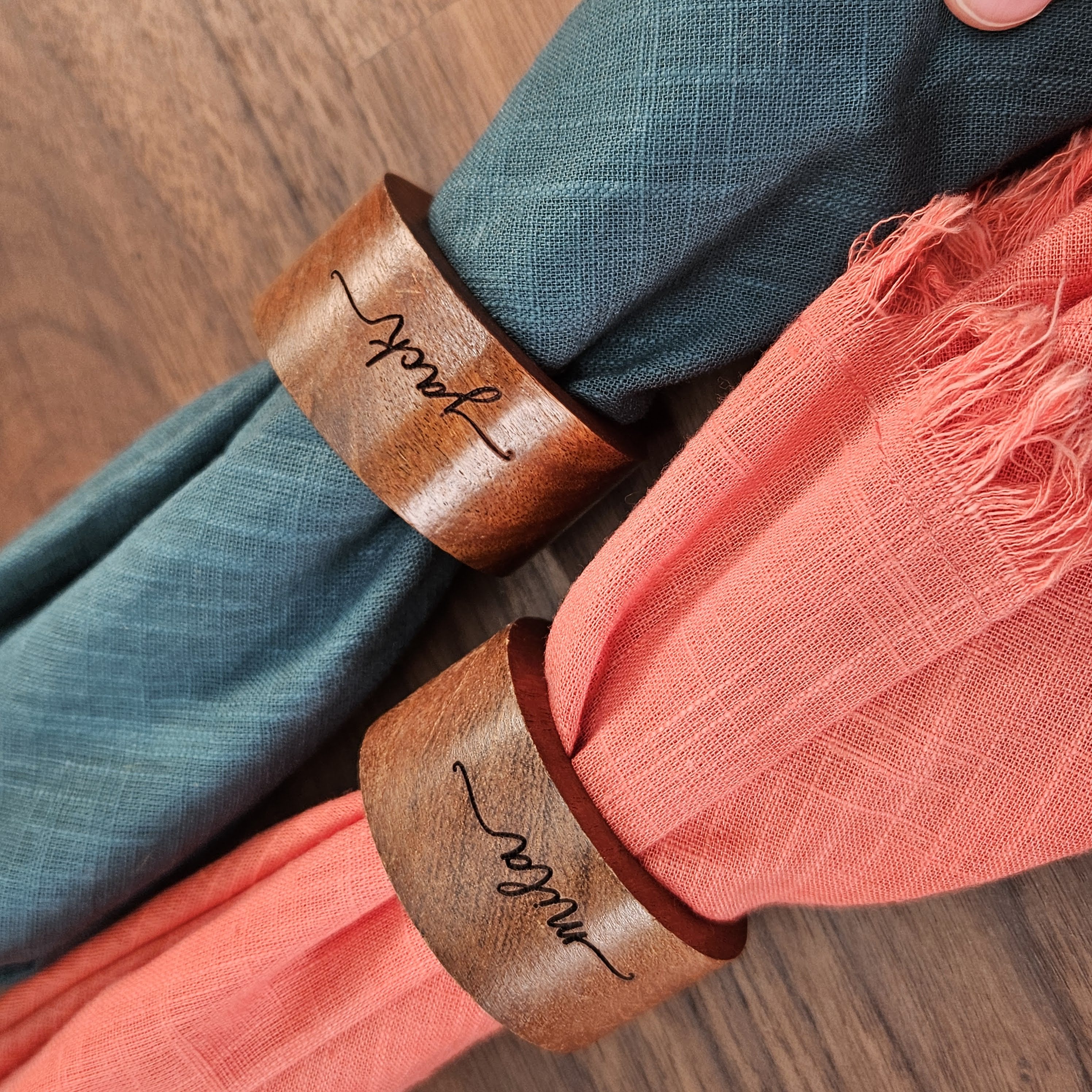 Custom laser engraved wood napkin rings personalized with names Mila and Jack, displayed on coral and teal napkins for a wedding or special event table setting. Made by Green Fern Studio, based in Seattle.