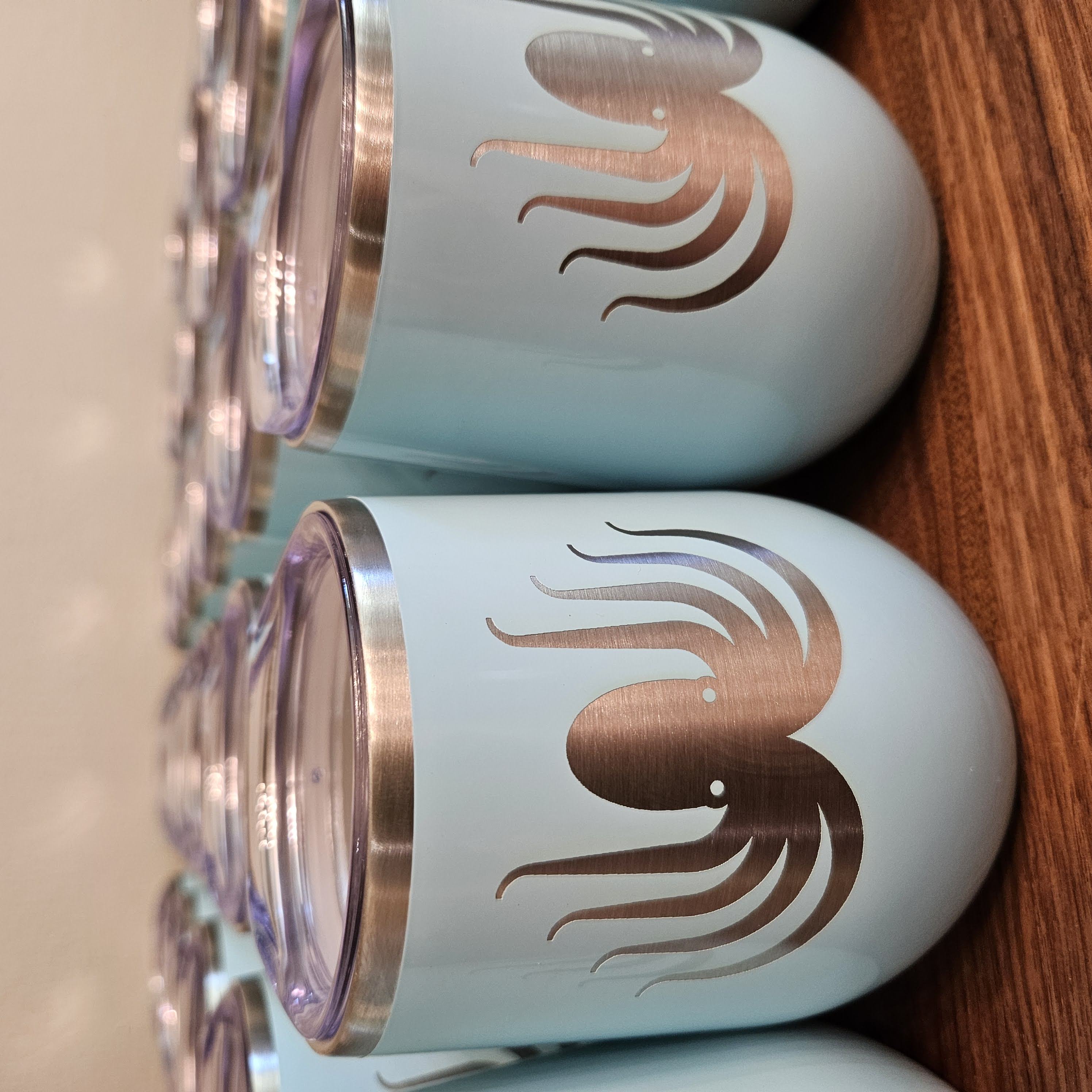 Light blue wine tumblers lined up on a wood surface with an octopus logo laser engraved on them