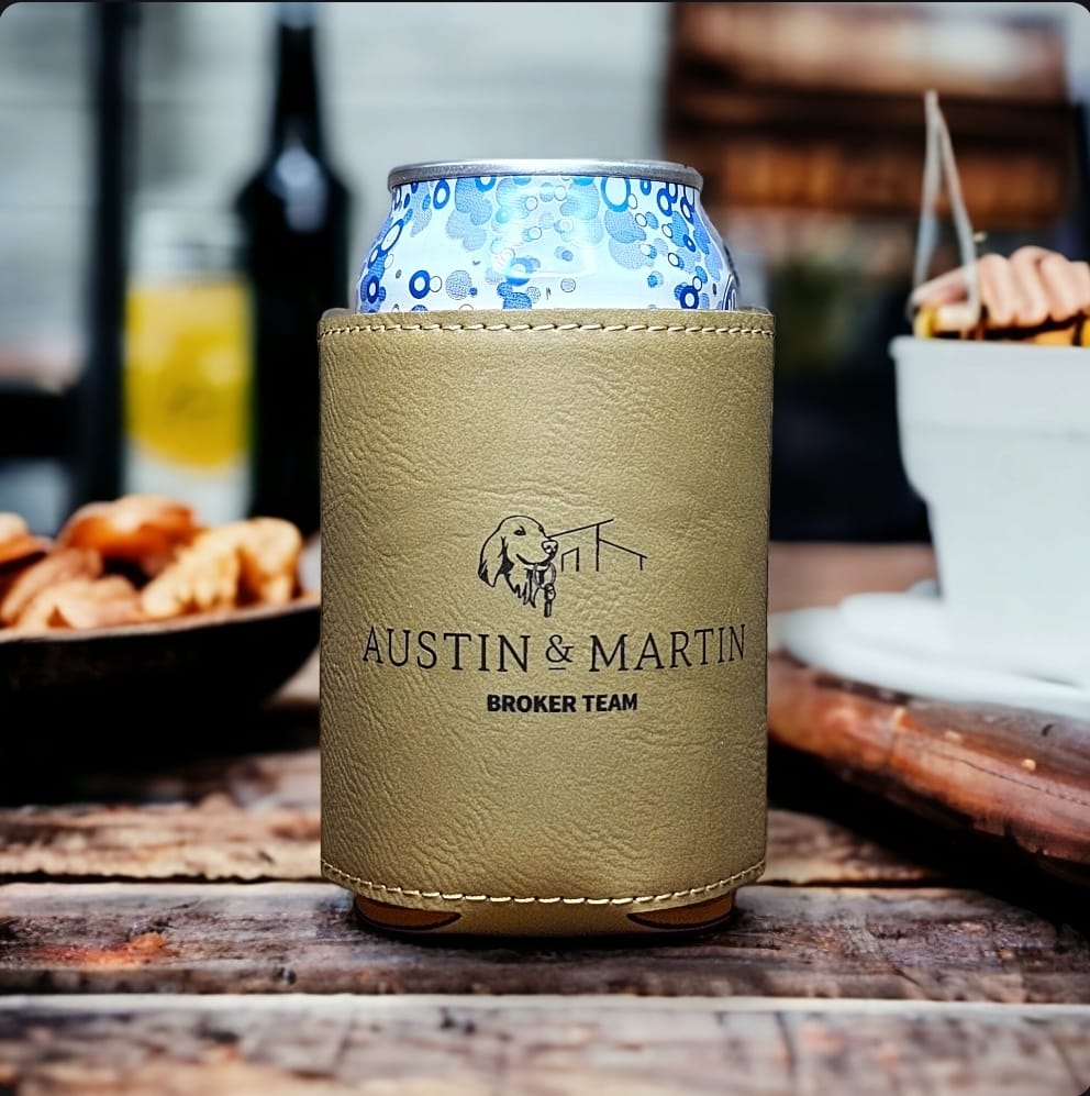Custom laser engraved soft vegan leather light brown koozie branded with Austin Martin Broker Team logo, displayed on a wood table. Made by Green Fern Studio, based in Seattle.