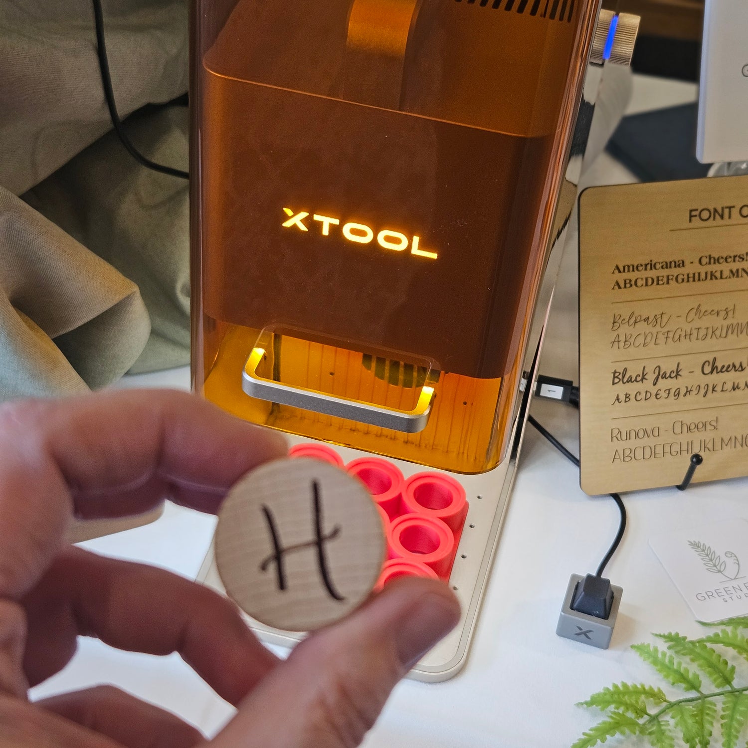 Hand holding a custom laser-engraved wood bottle stopper with the letter "H". An orange XTOOL laser engraving machine in the background.  Made by Green Fern Studio, based in Seattle.