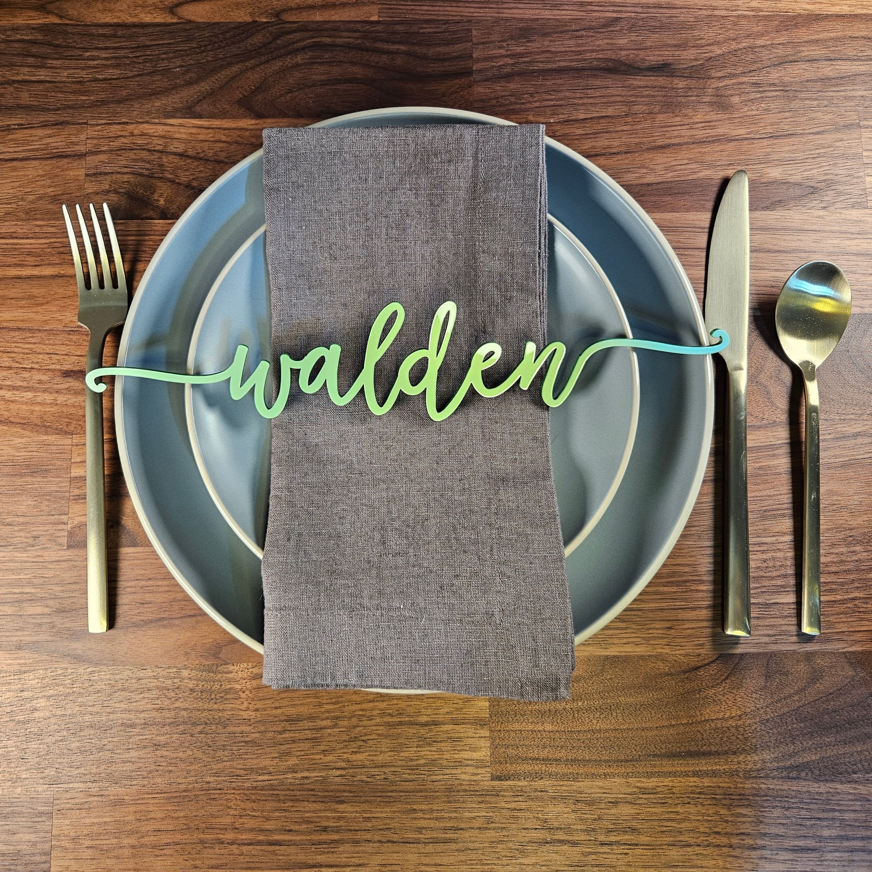 Jade green laser cut acrylic place setting engraved with the name Walden, elegantly displayed across grey plates with gold utensils on a warm wood table. Made by Green Fern Studio, based in Seattle.